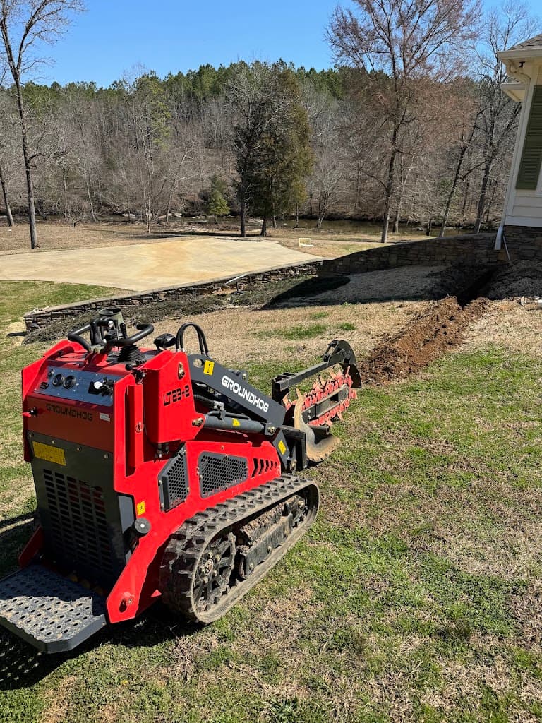 Mini skid-steer trencher digging a drainage line beside a house